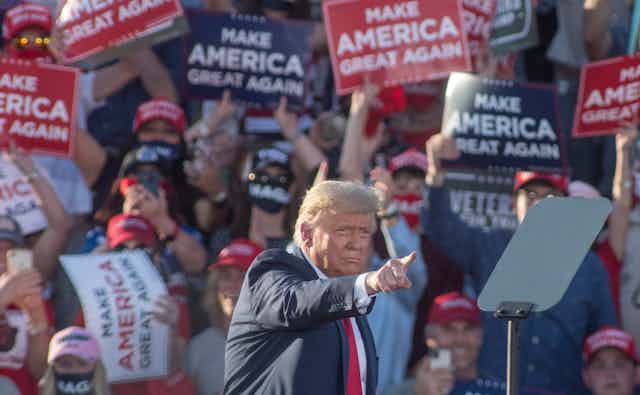Donald Trump pointing with supporters behind holding 'Make America great again' posters. 