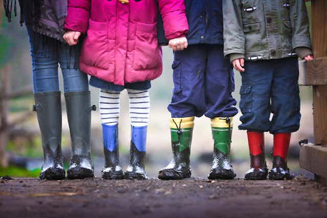Children in rainboots are seen from the neck down standing in a row.