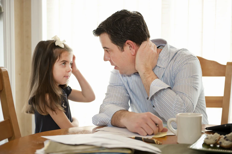 A man looks into the eyes of a little girl at the breakfast table while doing paperwork.