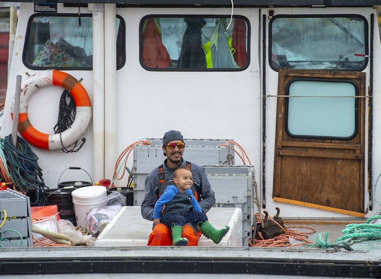 A man sitting at the back of a fishing boat with a smiling toddler on his lap.