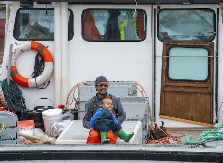 A man sitting at the back of a fishing boat with a smiling toddler on his lap.