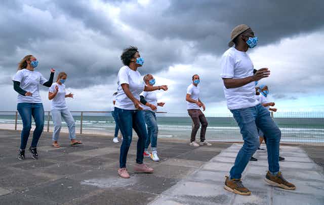 Eight men and women dance on a paved area near the sea, heavy clouds in the background. They are dressed in white T-shirts and jeans and wear matching face masks.