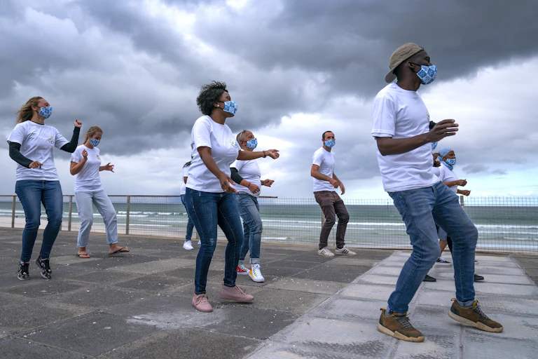 Eight men and women dance on a paved area near the sea, heavy clouds in the background. They are dressed in white T-shirts and jeans and wear matching face masks.