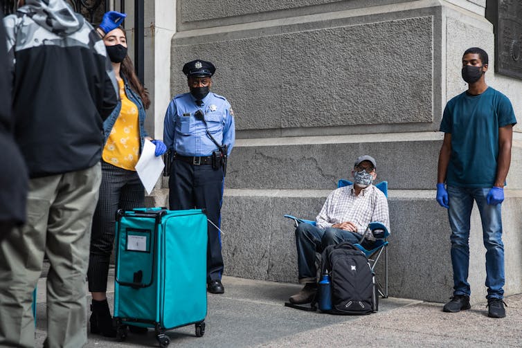 Man sits in folding chair near a line of voters and a police officer