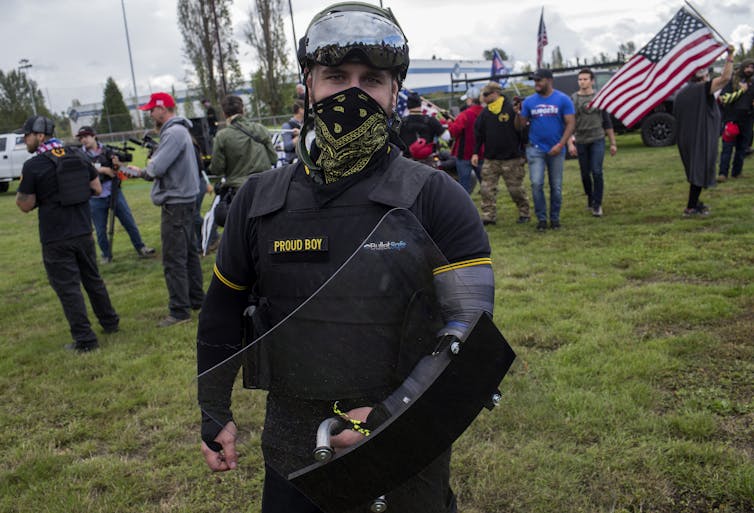 Picture of a man wearing military gear and the name 'Proud Boys' with an American flag in background
