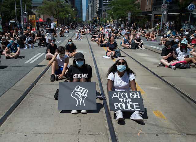 Protesters sit on a road with signs showing a Black power fist and ABOLISH THE POLICE