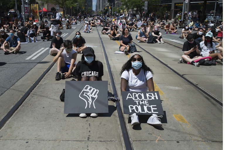 Protesters sit on a road with signs showing a Black power fist and ABOLISH THE POLICE