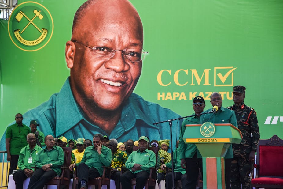 A portrait of s smiling President John Magufuli, wearing specs, looms large over seated party officials as he speaks from a podium, flanked by a solder military fatigue on the right, and male party official in a green shirt, on the left