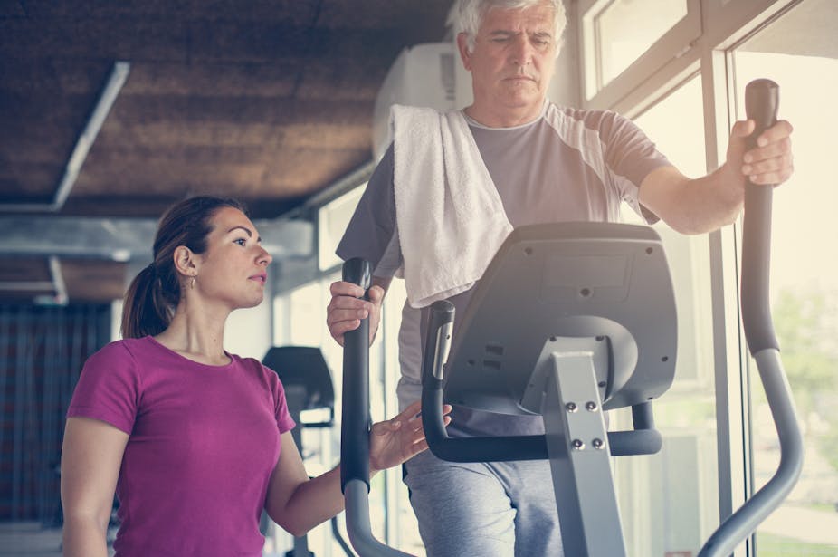 Older man exercises on elliptical with personal trainer beside him.