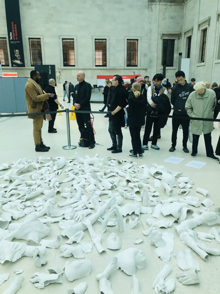 Observers behind a fence look at artwork of white faces and arms in foreground.