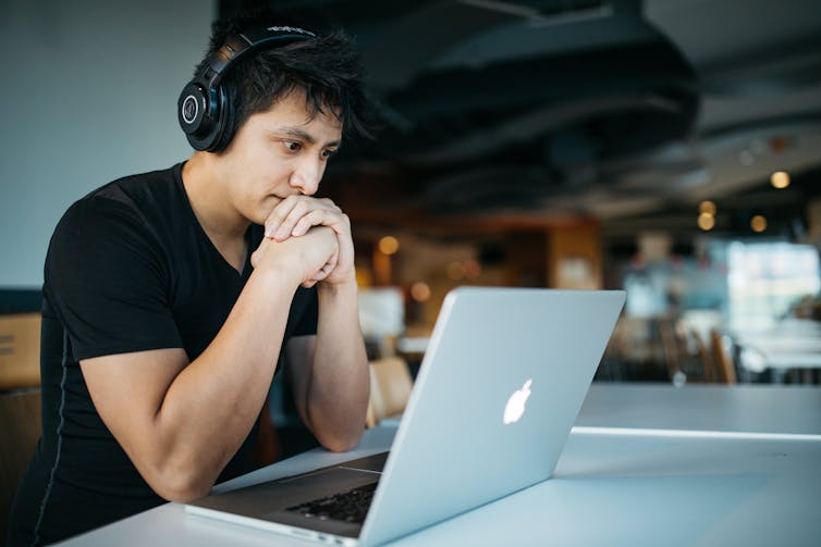 A man wearing earphones looks at his laptop screen.