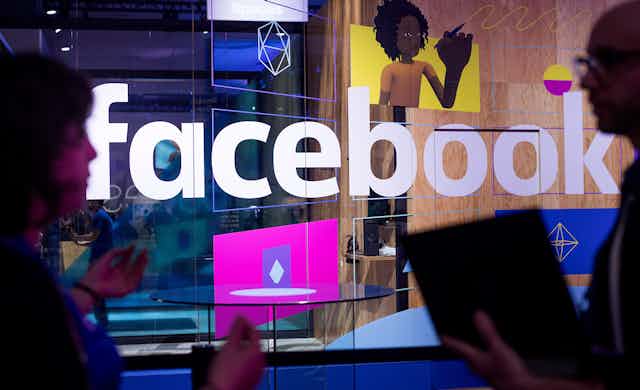 A man and a woman speaking in front of a glass wall containing the Facebook logo