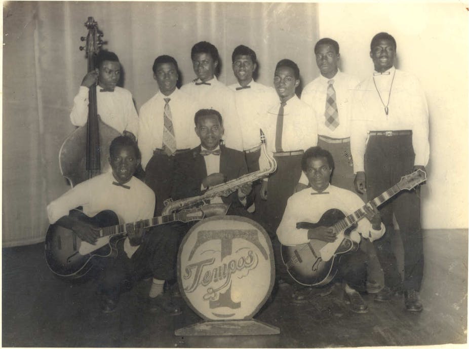 Men in white shirts and dark trousers, some holding musical instruments, pose for a group photo, with the name of the band displayed in front of the group