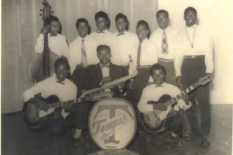 Men in white shirts and dark trousers, some holding musical instruments, pose for a group photo, with the name of the band displayed in front of the group