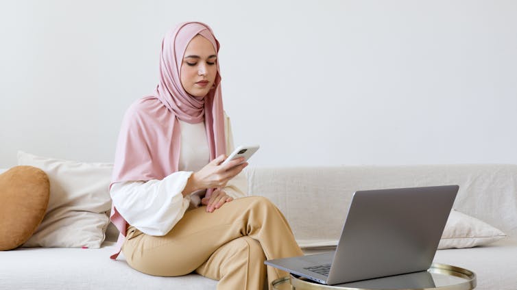 A woman in a pink head scarf looks at her phone with her laptop in front of her.