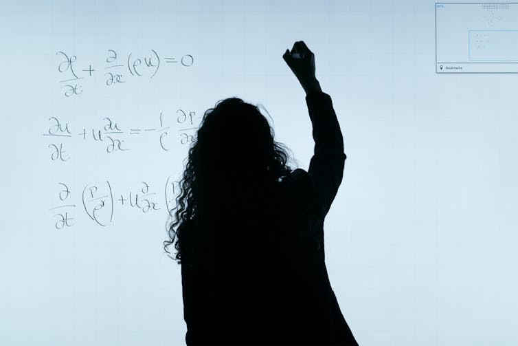 A woman is silhouetted in front of a white board as she writes mathematical equations on it.