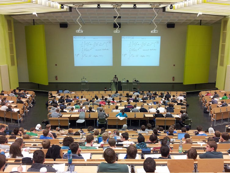 A lecture hall is seen filled with students and a professor at a podium.