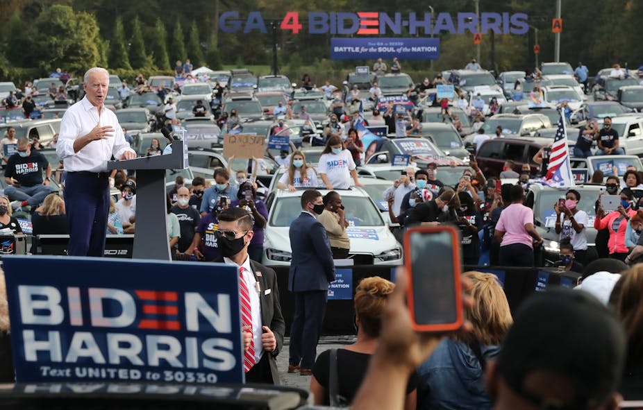 Joe Biden standing in front of a lectern with people watching by their cars at a rally.