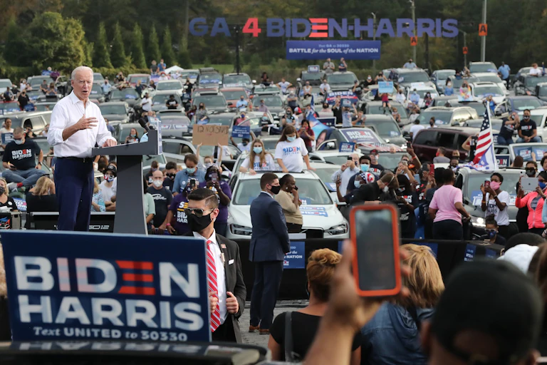 Joe Biden standing in front of a lectern with people watching by their cars at a rally.