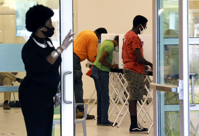 Woman stands by door of room where people are voting using machines.