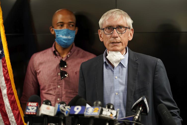 Wisconsin Gov. Tony Evers standing at a podium speaking with a face mask pulled down around his neck. Behind him is a man wearing a face mask.