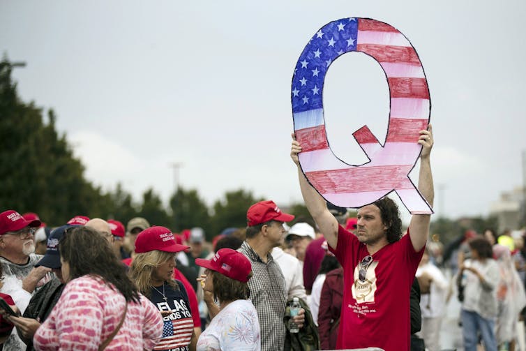 A man in a red T-shirt holds up a letter Q illustrated with stars and stripes.