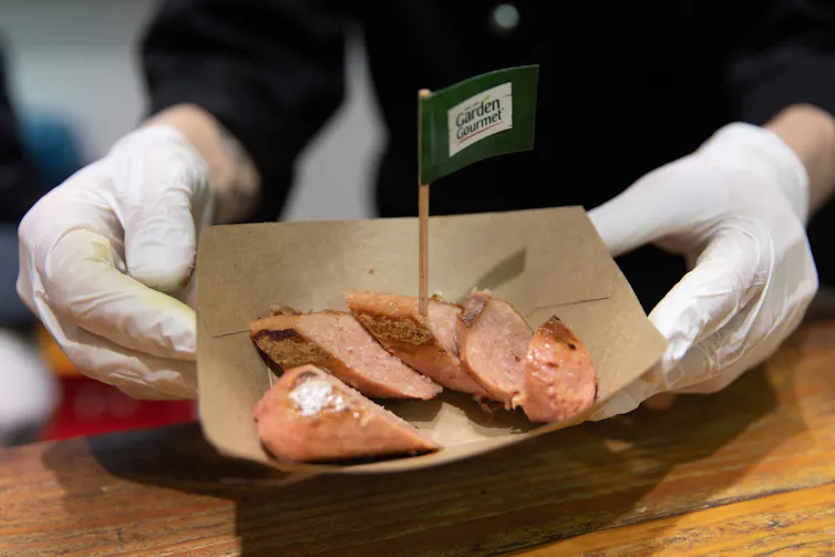 A pair of gloved hands hold a cardboard tray of plant-based sausage samples.
