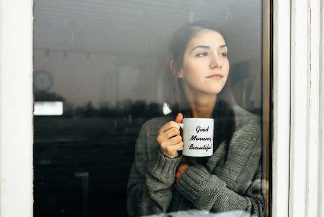 A woman holding a mug saying 'good morning beauitful' looks out of a window. 