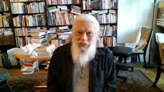 An older man with a huge white beard sits looking at camera, behind him a study and shelves of books.