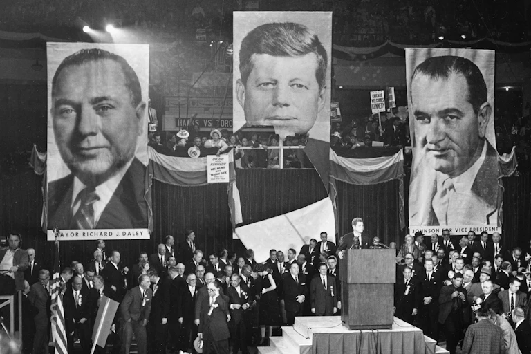 Presidential candidate John F. Kennedy speaks before a throng of supporters in front of banners depicting the faces of Kennedy, Chicago mayor Richard Daley and vice presidential candidate Lyndon B. Johnson.