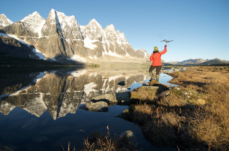 A person holding an antenna in the air near a lake with a backdrop of snow peaked mountains.