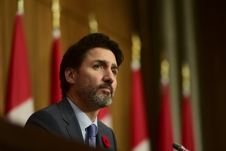 A bearded Trudeau is seen at a news conference with flags behind him.