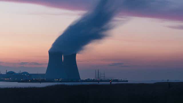 Twin cooling towers release steam at dusk with a bay in the foreground.