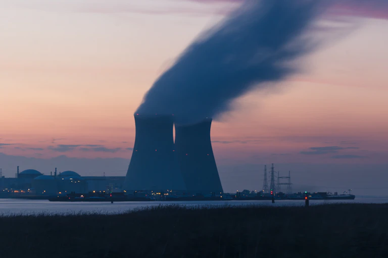 Twin cooling towers release steam at dusk with a bay in the foreground.