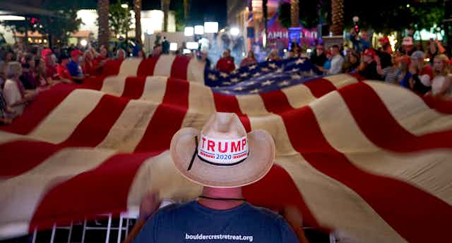 A person wearing a cowboy hat with a Trump sticker standing at the end of a large American flat.