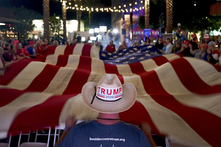 A person wearing a cowboy hat with a Trump sticker standing at the end of a large American flat.