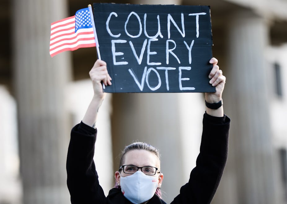 Woman holds up a sign saying 'count every vote' with a US flag. 