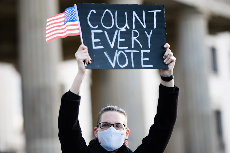 Woman holds up a sign saying 'count every vote' with a US flag.