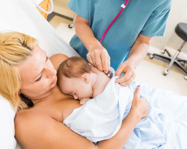 A woman holds her infant in skin-to-skin contact while a health-care professional holds a stethoscope to the baby's back.