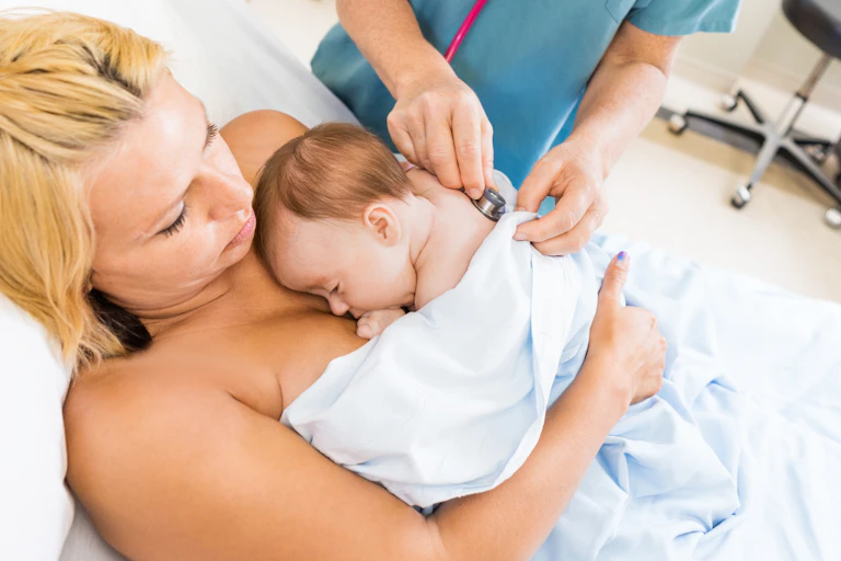 A woman holds her infant in skin-to-skin contact while a health-care professional holds a stethoscope to the baby's back.