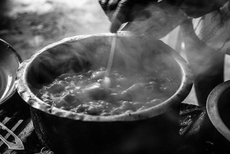 A hand stirs a steaming and battered metal pot with a spoon. The contents resemble a stew.