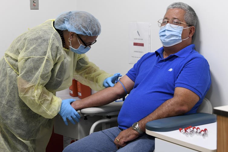 A man wearing a face mask with a tourniquet around his arm sits in a chair beside some empty vials while a woman wearing personal protective equipment looks for a vein in his arm.