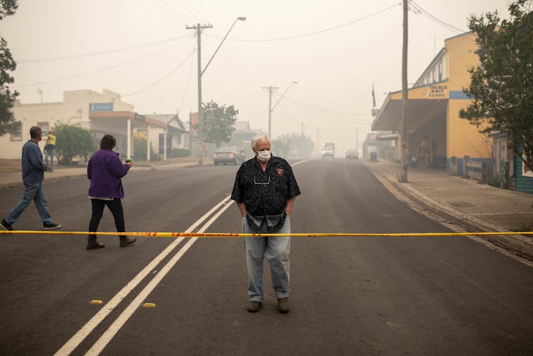 A man standing behind a cordoned off area with thick smoke behind him, in Cobargo, NSW.