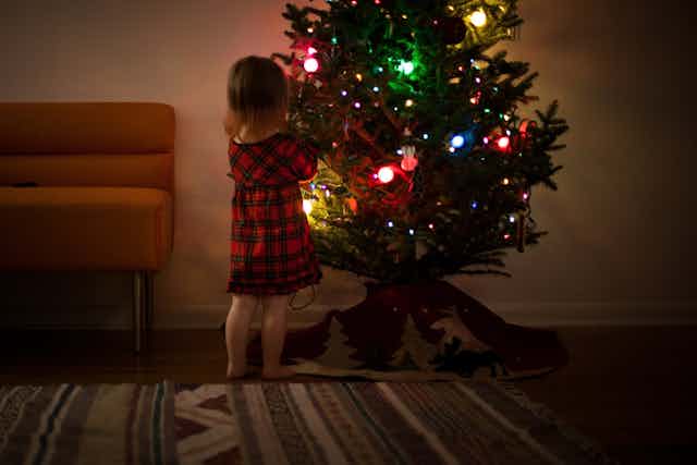 Little girl in tartan dress looks at Christmas tree in living room.