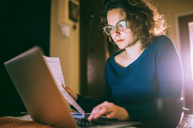 A girl holds a piece of paper while working on a laptop.