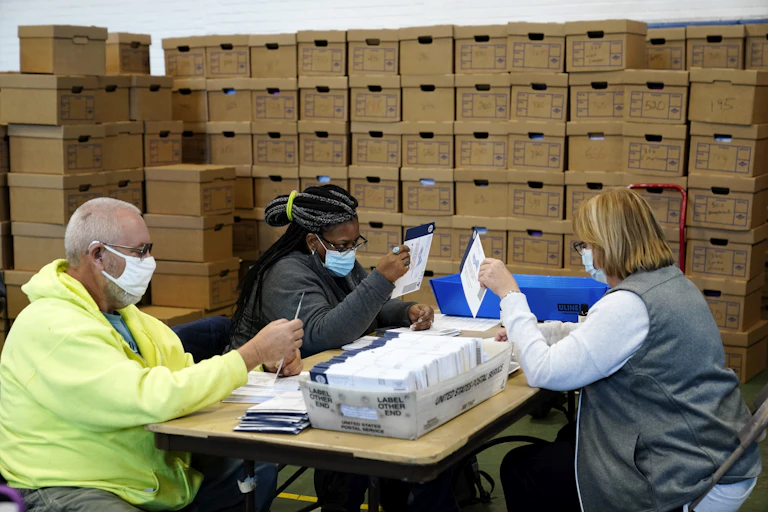 A man and two women look at envelopes with a been similar envelopes on a table between them and stacks of cardboard boxes behind them