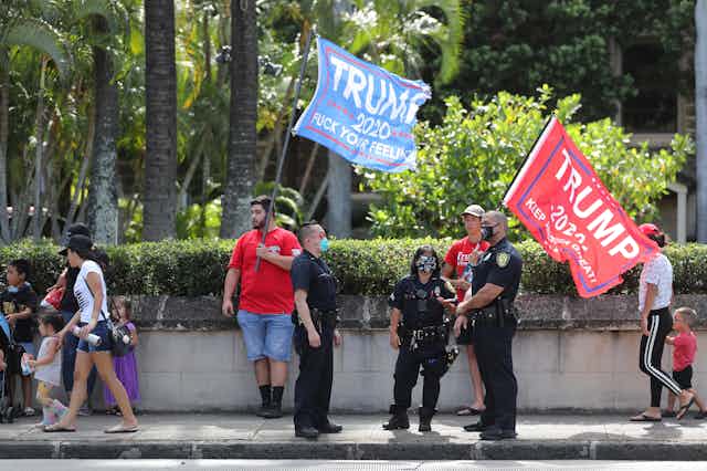 Three police officers stand in front of Trump supporters carrying Trump 2020 flags