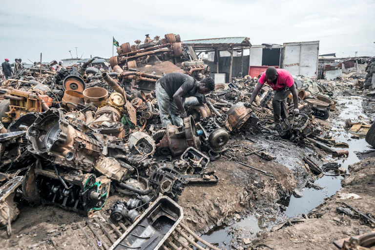Two men sort through heap of discarded metal items next to a ditch