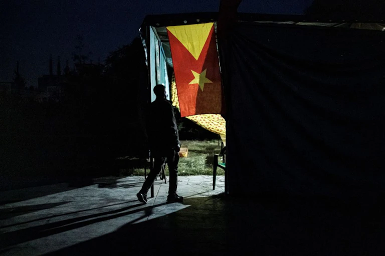 a man stepping from darkness towards a door with light shining inside and a red and yellow flag with a triangle and a star hanging inside.