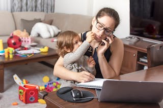 Young mother holding child as she tries to speak on the phone while working from home.